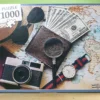 Das "Time to Travel" 1000 Teile Puzzle von vorn. Es zeigt ein Portemonnaie mit Geldscheinen, eine alte Fotokamera, eine Uhr, einen Strohhut und eine Sonnenbrille, alles auf einer Weltkarte platziert.