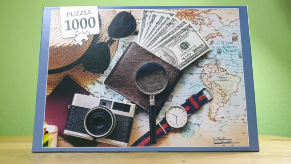 Das "Time to Travel" 1000 Teile Puzzle von vorn. Es zeigt ein Portemonnaie mit Geldscheinen, eine alte Fotokamera, eine Uhr, einen Strohhut und eine Sonnenbrille, alles auf einer Weltkarte platziert.